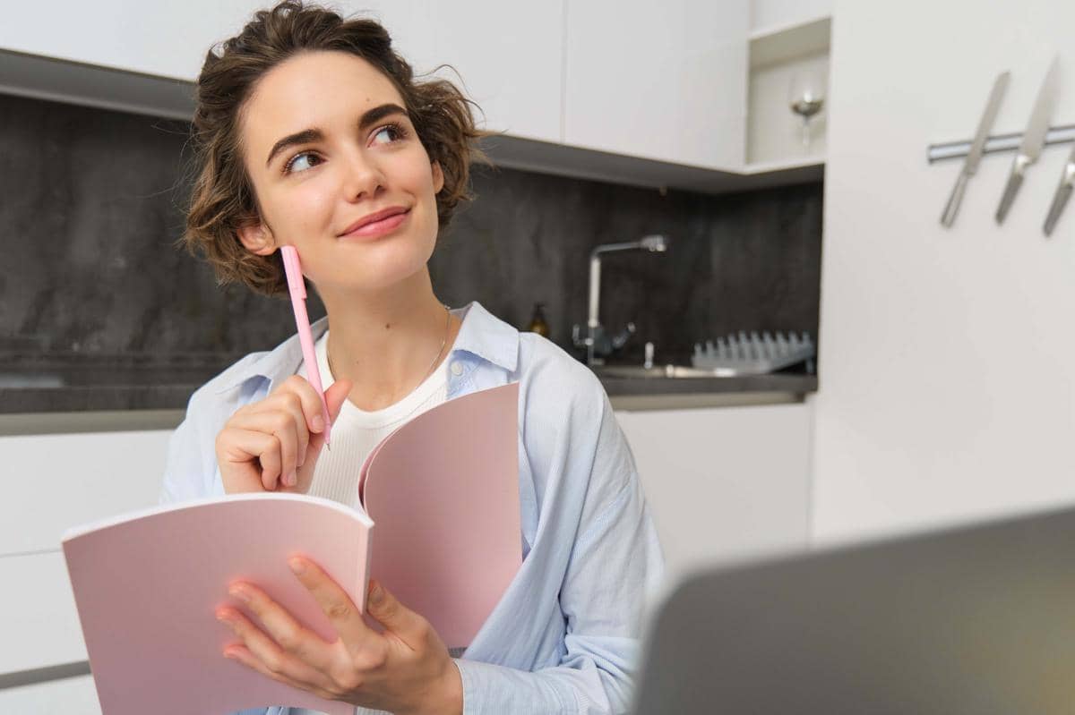 Seorang perempuan muda duduk di rumah sambil menulis catatan pada buku perencana di atas meja dengan ekspresi berpikir.