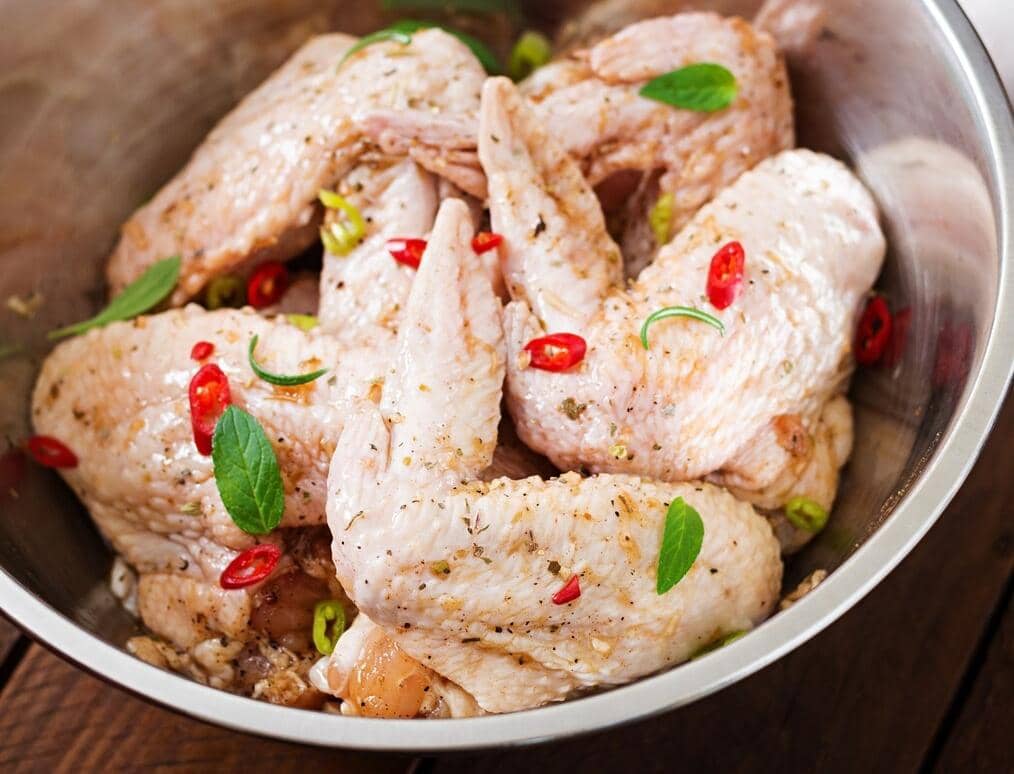 Potongan ayam mentah dalam mangkuk putih diberi bumbu marinasi, dikelilingi bawang putih, cabai, dan rempah.