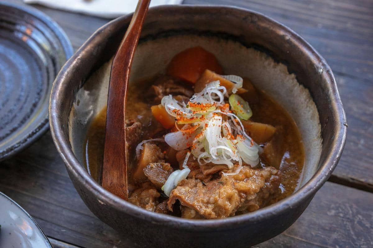 Ilustrasi semangkuk chamchi jjigae berkuah merah dengan tahu, sayuran, dan nasi putih hangat di meja kayu.