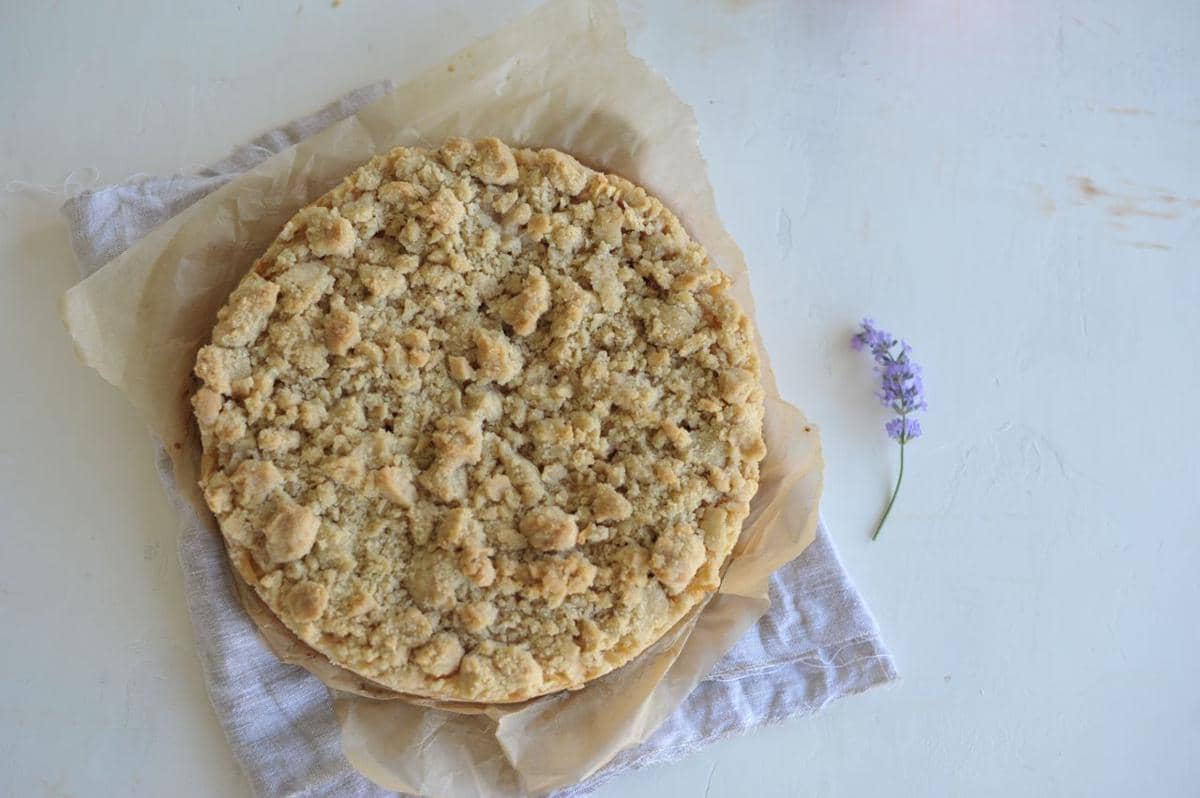Sepotong apple crumble cake berlapis remah renyah disajikan di atas piring, dengan irisan apel sebagai pelengkap.