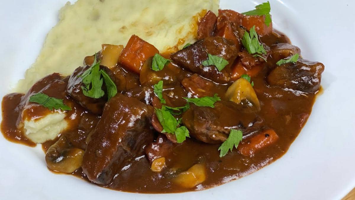 Ilustrasi semangkuk beef bourguignon berisi potongan daging sapi dalam kuah cokelat, disajikan dengan kentang dan sayuran.