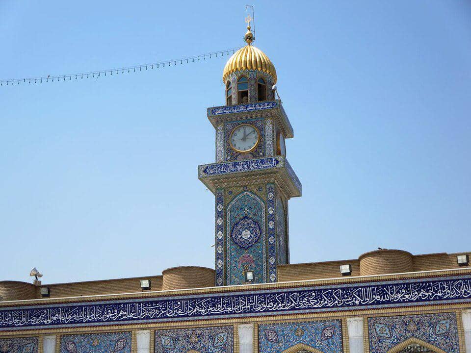 Menara jam Masjid Agung Kufa menjulang dengan kubah kecil dan ornamen arsitektur Islam di bawah langit cerah.