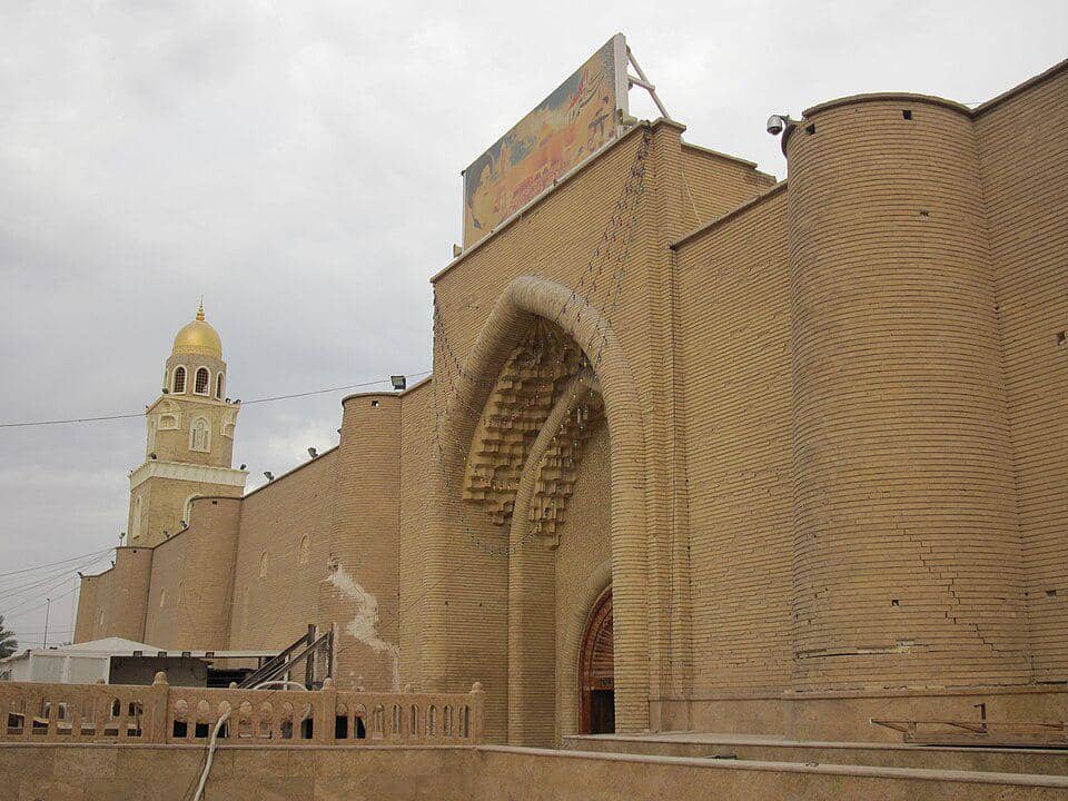 Pintu masuk Masjid Agung Kufa di Irak dengan lengkungan bernuansa biru, kaligrafi Arab, dan ornamen Islam.