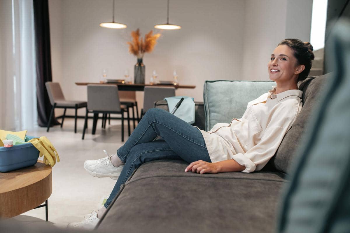 Seorang perempuan muda tersenyum sambil duduk santai di sofa ruang keluarga, menggambarkan suasana rumah yang nyaman.