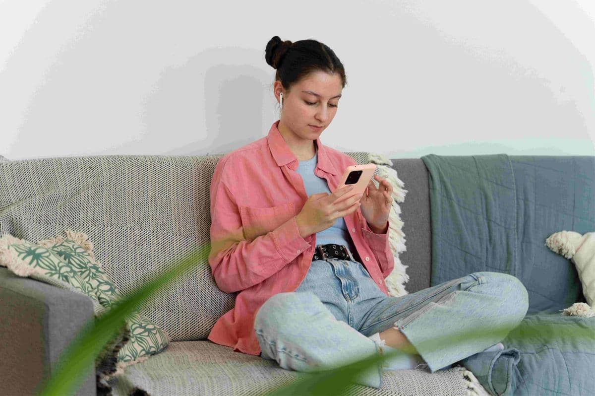 Seorang perempuan duduk santai di sofa sambil menatap dan menggulir layar ponsel pintar di dalam ruangan.