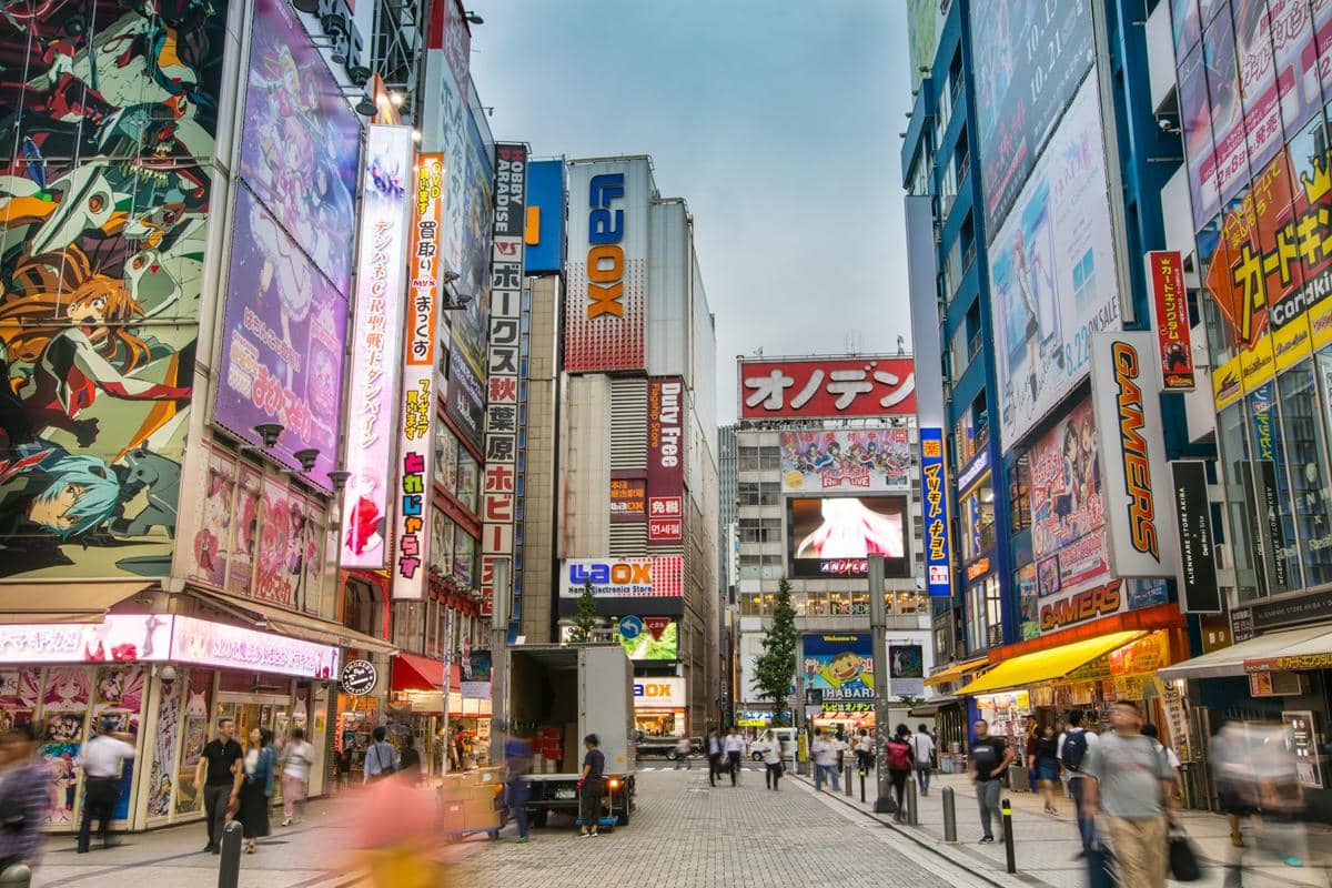 Jalan di Akihabara, Jepang, dipenuhi gedung bertingkat, papan reklame bercahaya, dan pejalan kaki pada malam hari.