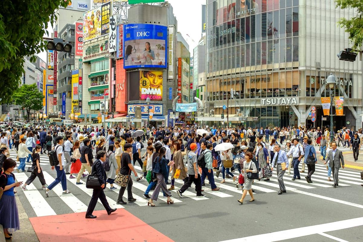 Suasana Shibuya Crossing di Jepang pada malam hari dengan pejalan kaki melintasi zebra cross dan gedung bercahaya.