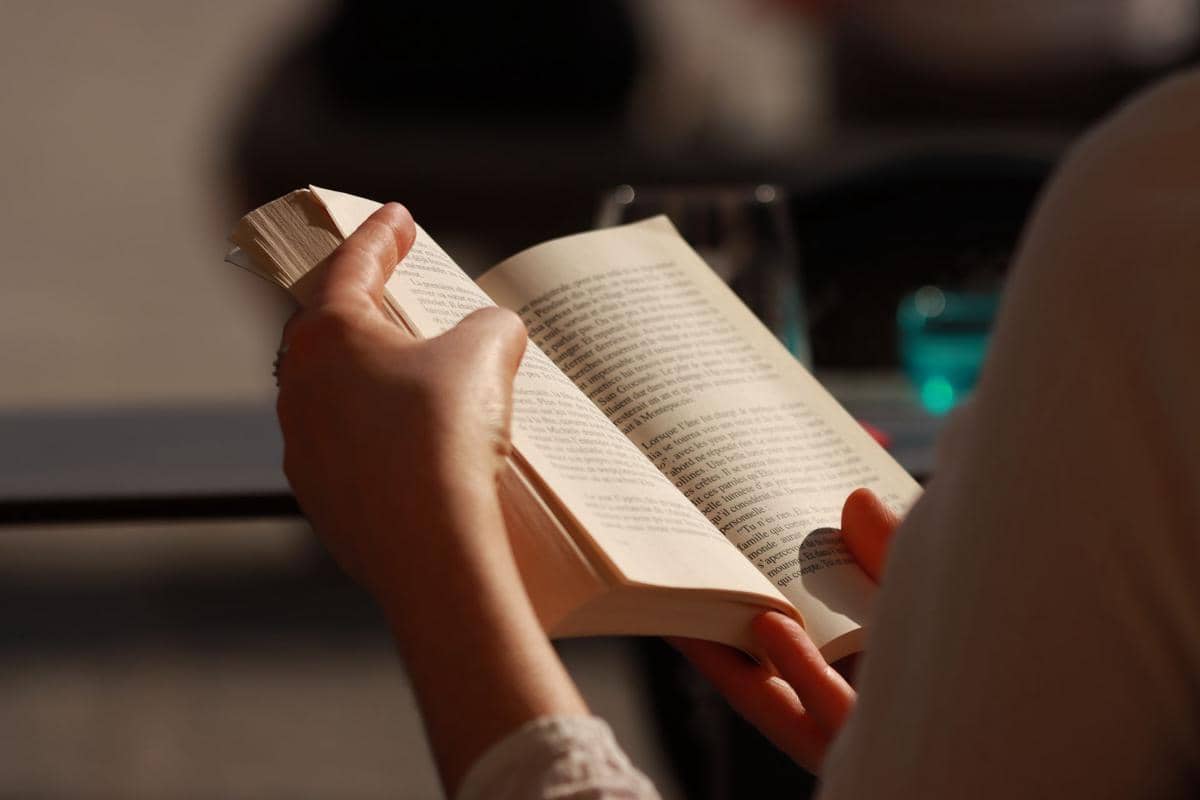 Tangan membuka halaman buku tebal di atas permukaan kayu, menggambarkan aktivitas membaca dan belajar.