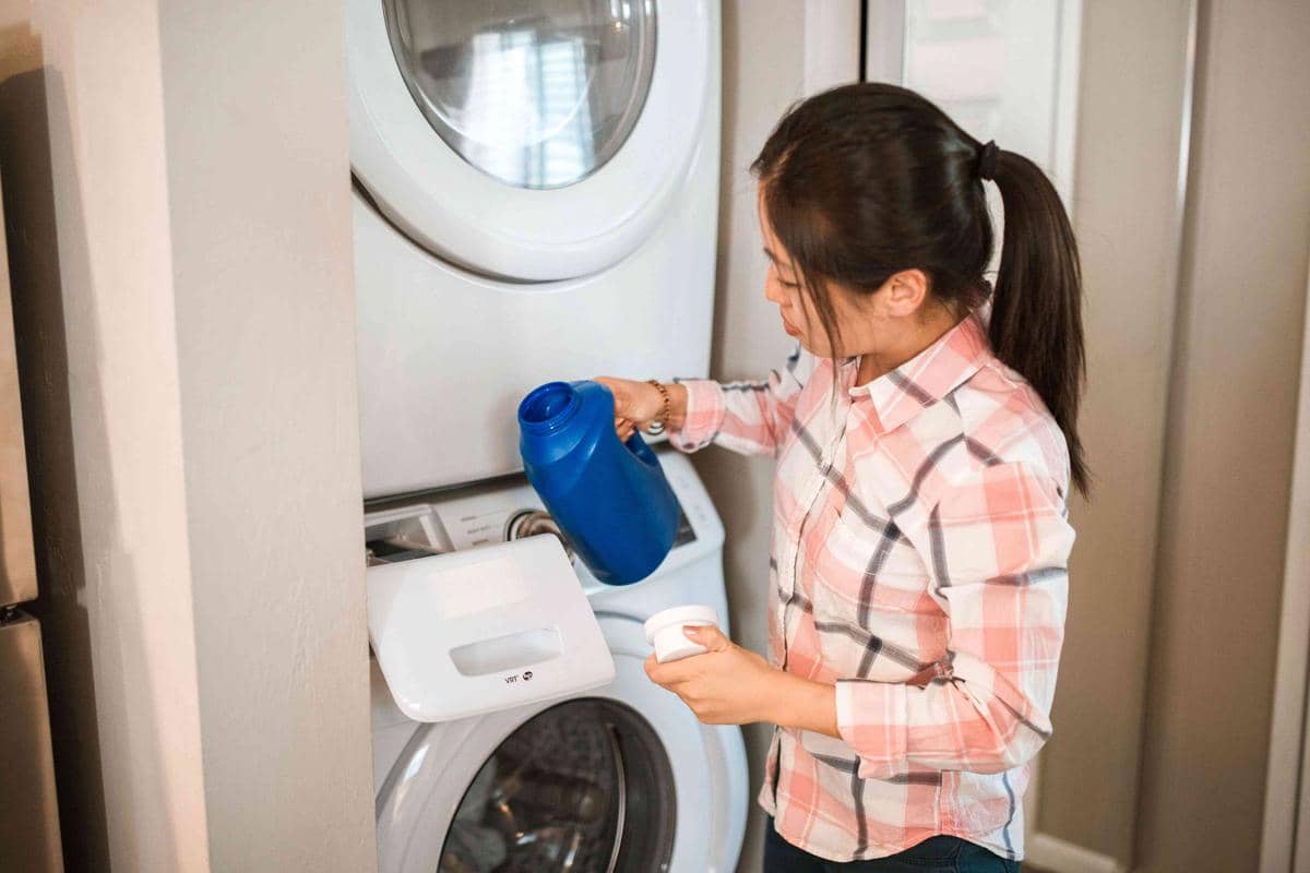 Ilustrasi seseorang mencuci pakaian di rumah menggunakan mesin cuci di area laundry dengan keranjang baju.