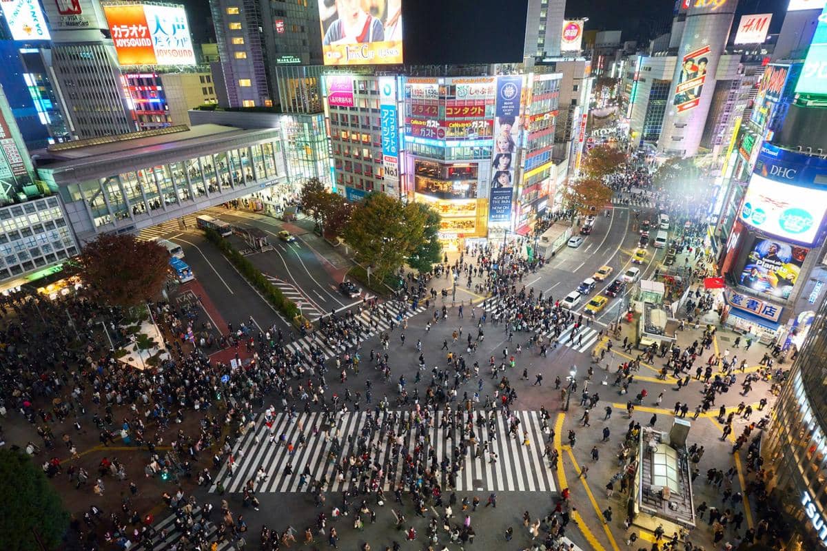 Pejalan kaki melintasi zebra cross di Shibuya Crossing, Tokyo, dengan gedung tinggi dan papan iklan bercahaya di sekelilingnya.