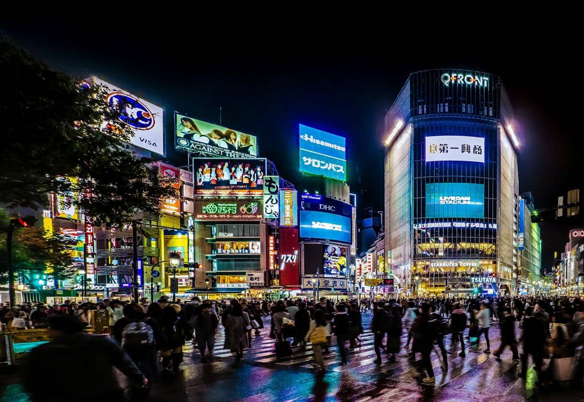 Pejalan kaki melintasi zebra cross di Shibuya Crossing, Tokyo, dengan gedung-gedung dan papan iklan di sekelilingnya.