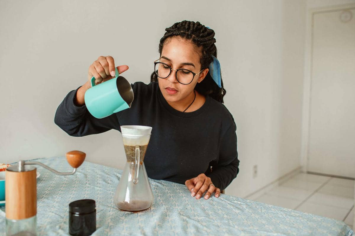 Tangan menuangkan air panas dari ketel ke dripper kopi dengan cangkir di atas meja kayu untuk proses seduh.