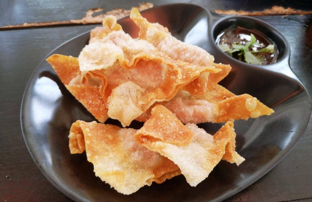 Sepiring pangsit goreng berwarna keemasan disajikan di atas piring putih, dengan beberapa potong tersusun bertumpuk.