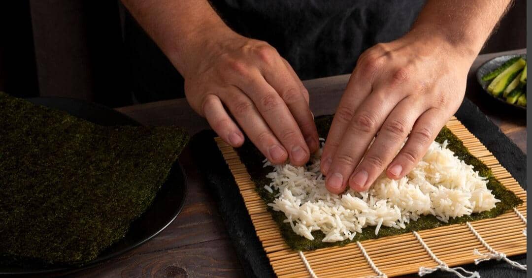 Tangan seorang koki menggulung nasi dan isian di atas lembaran nori dalam proses pembuatan sushi di dapur.