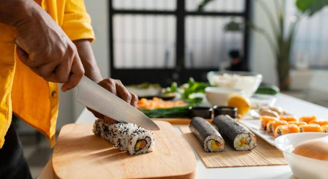 Tangan menggunakan pisau tajam untuk memotong gulungan sushi di atas talenan kayu, menghasilkan beberapa potongan rapi.