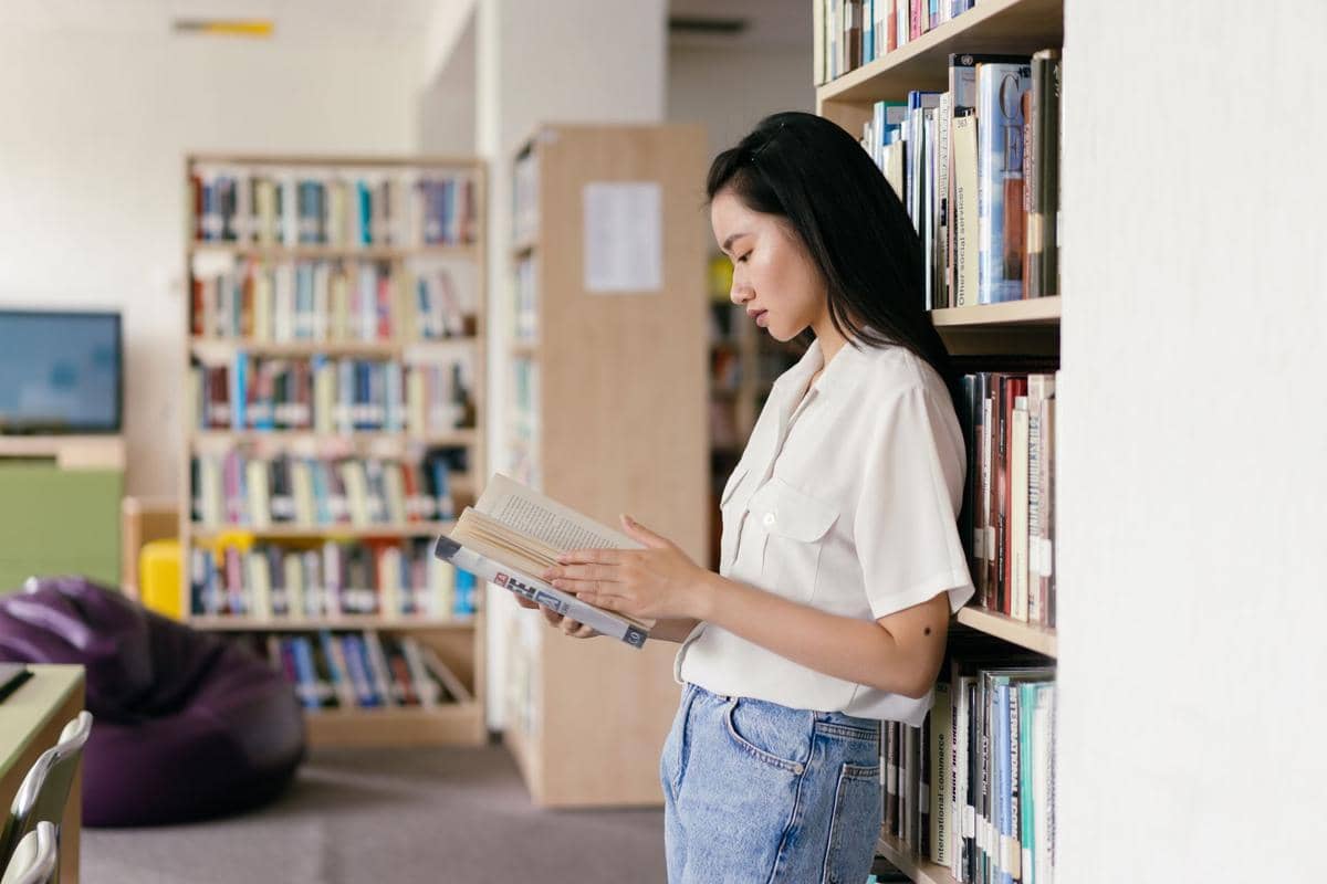 seorang wanita membaca buku sambil bersender di rak buku