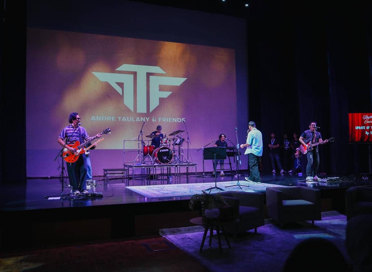 Andre Taulany dan Friends menghadiri konser amal sekolah Kenzy, berdiri bersama di panggung acara yang dihiasi layar besar.