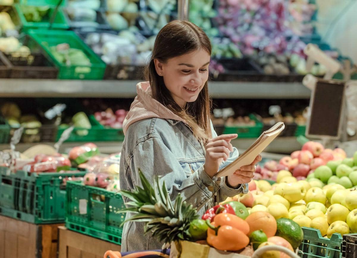Seorang perempuan muda memeriksa daftar belanja di buku catatan sambil memilih bahan makanan di supermarket.