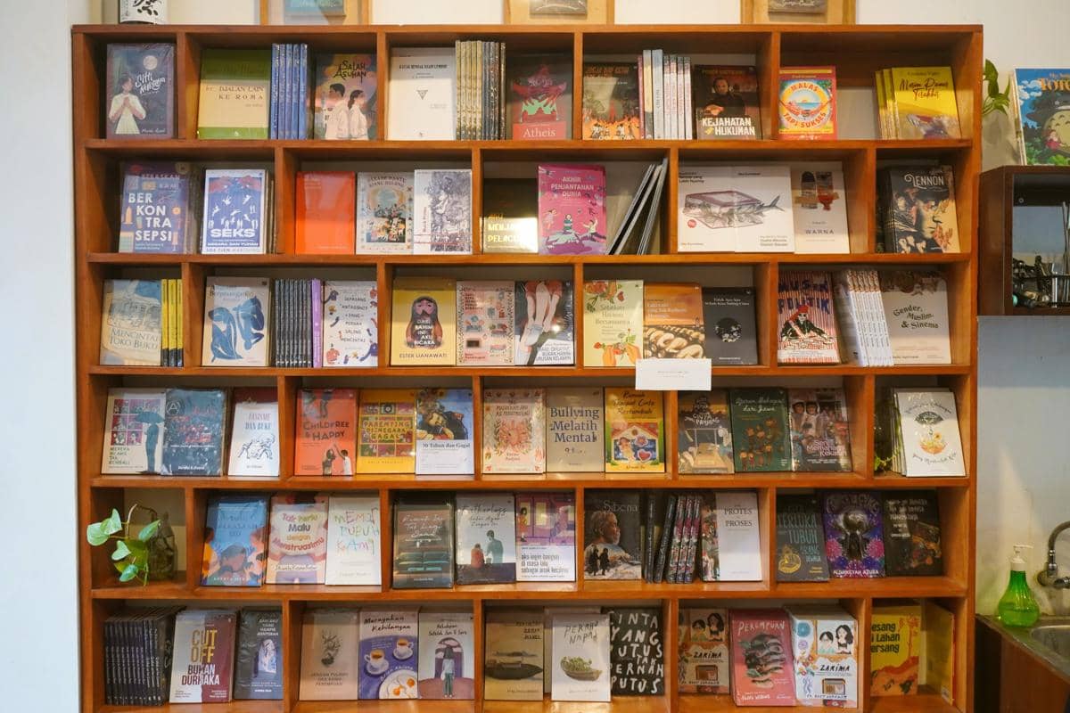 Rak buku kayu berisi deretan buku dengan sampul beragam tersusun rapi, menciptakan suasana ruang baca yang hangat.