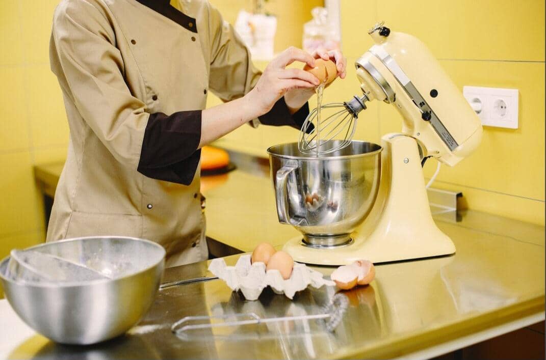 Tangan memasukkan telur ke dalam mangkuk mixer bersama bahan-bahan untuk membuat adonan di atas meja dapur.