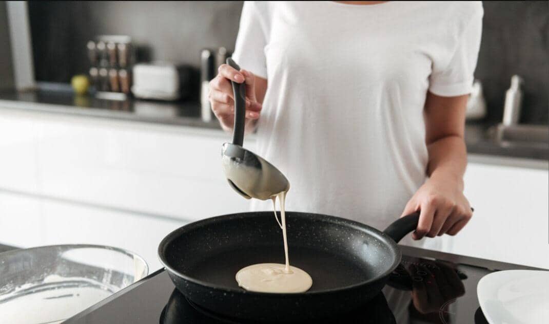 Tangan menuang adonan pancake dari wadah ke wajan teflon panas di dapur untuk dimasak hingga benar-benar matang.