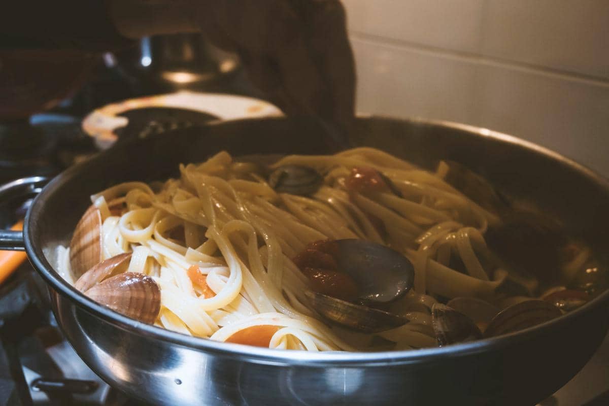 Semangkuk pasta berbentuk kerang disajikan dengan saus tomat, keju parut, dan daun basil di atas meja.