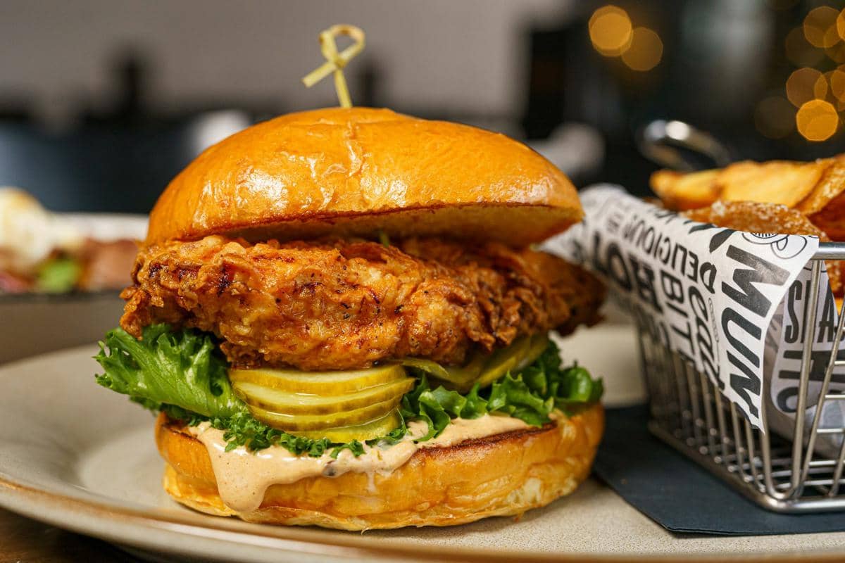 Sandwich ayam lada hitam dengan roti panggang, selada, tomat, dan saus yang disajikan di atas piring.