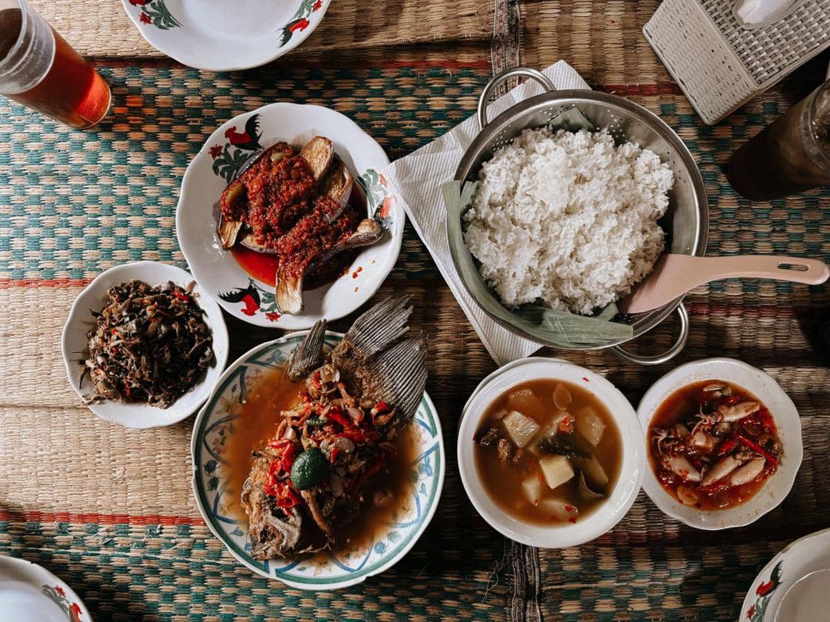Sepiring nasi putih dengan aneka lauk dan makanan pendamping tersaji di atas meja makan bersama beberapa hidangan lain.