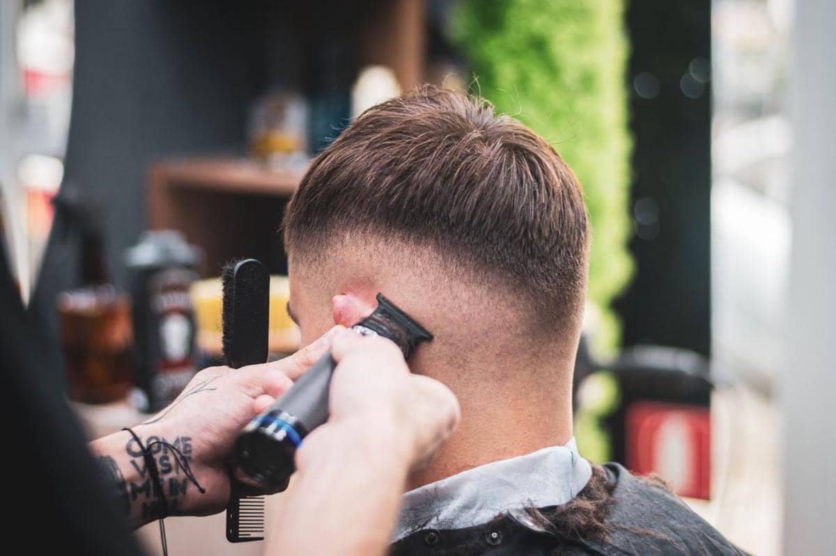 Seorang pria duduk di kursi barbershop saat rambutnya dipotong oleh penata rambut menggunakan alat cukur.