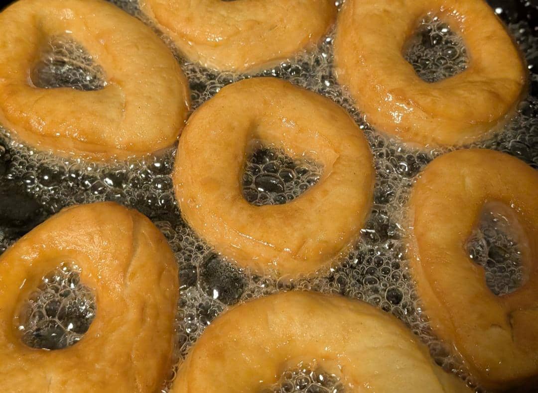 Tangan menggunakan penjepit membalik beberapa donat yang digoreng dalam wajan berisi minyak panas mendidih.