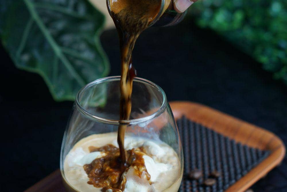 Tangan menuangkan espresso dari gelas kecil ke dalam cangkir berisi susu, membentuk lapisan kopi untuk minuman latte.