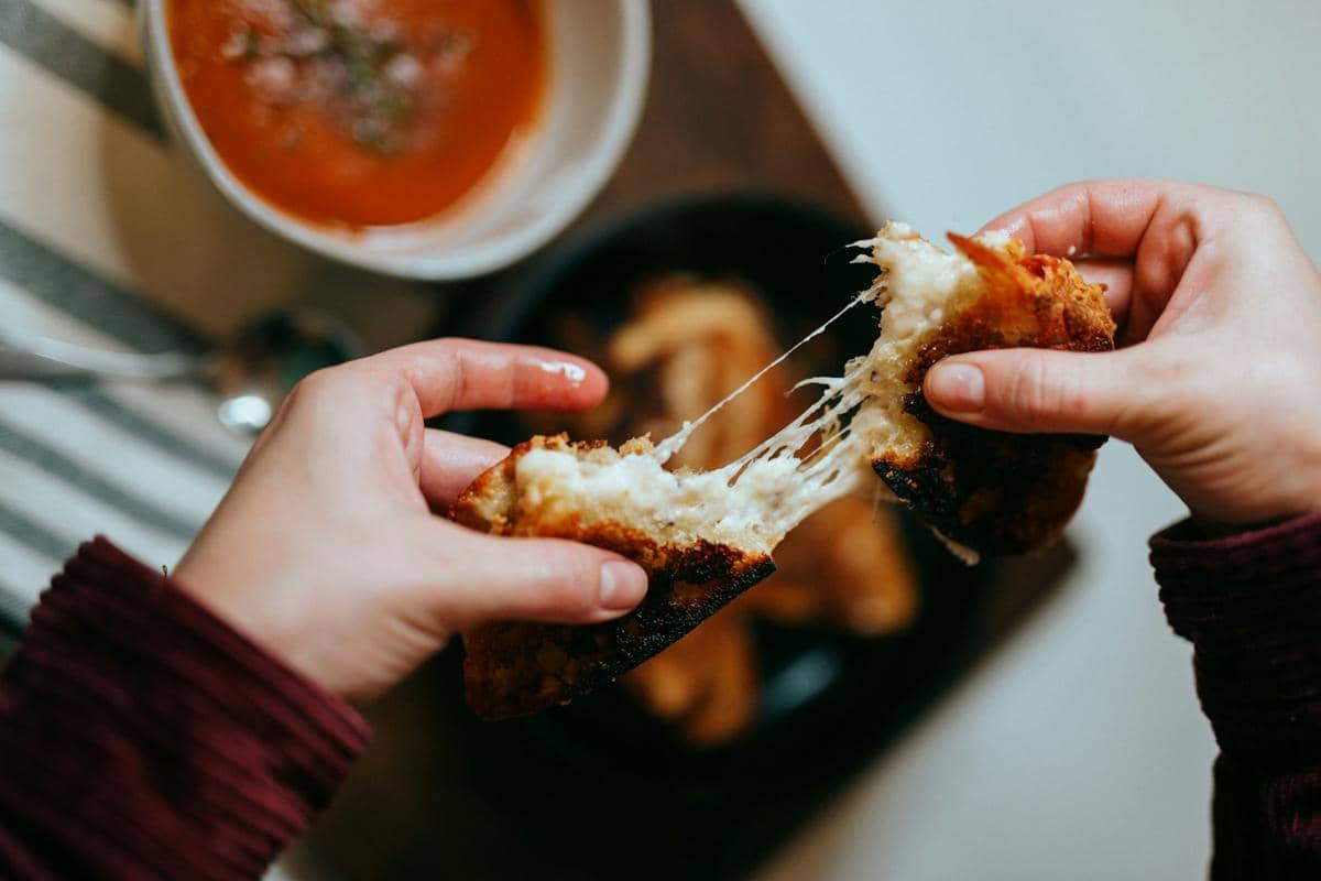 Semangkuk sup tomat hangat disajikan bersama potongan roti panggang keju di atas meja kayu bernuansa gelap.