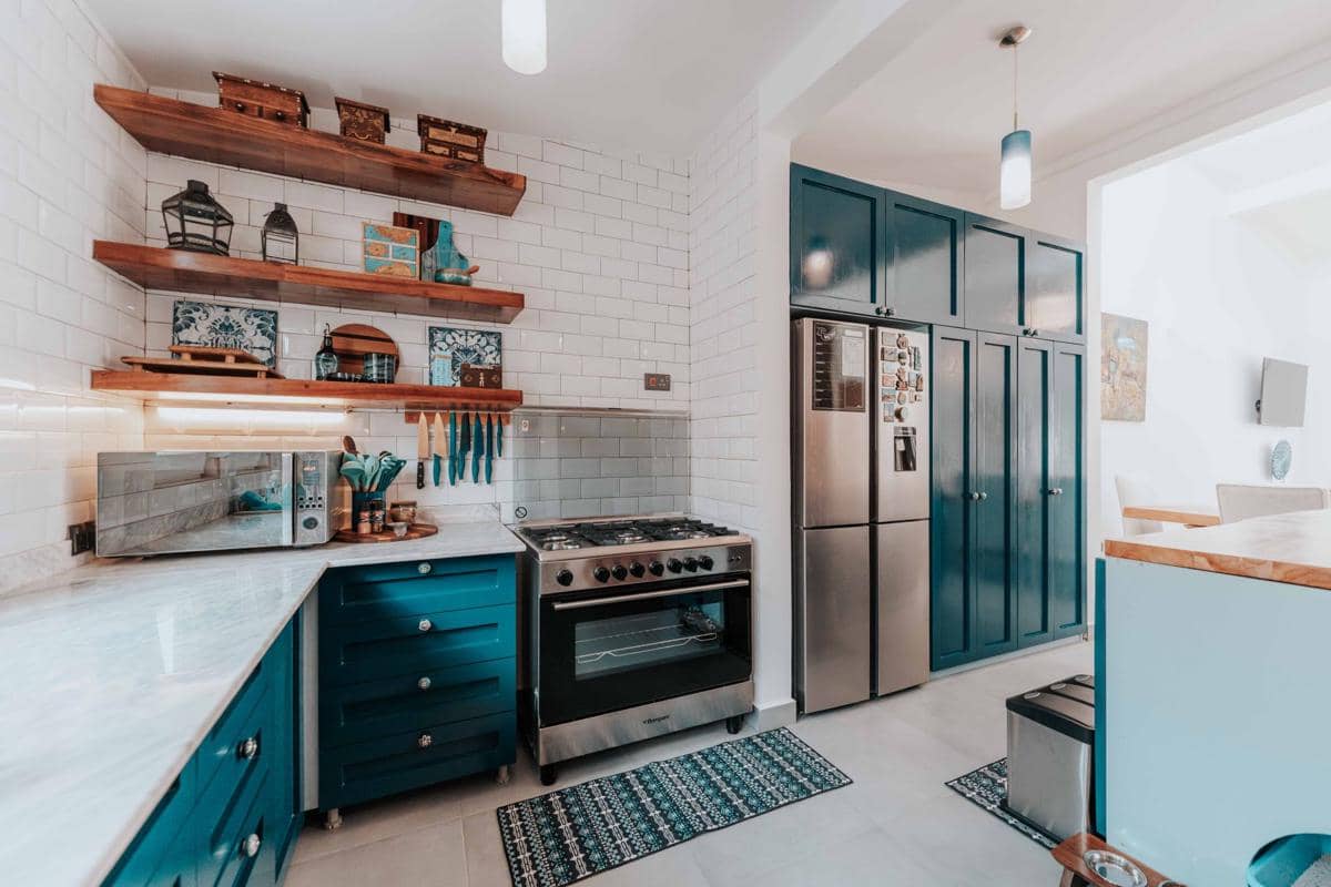 Ilustrasi dapur modern dengan kulkas putih, meja, rak, dan peralatan rumah tangga dalam pencahayaan hangat.