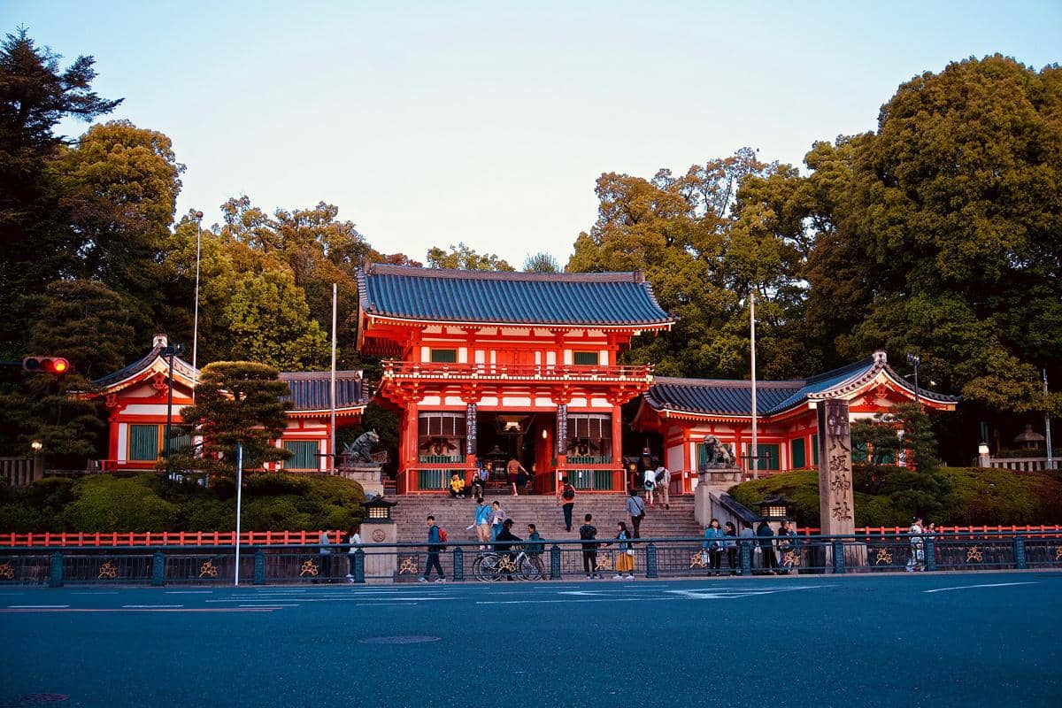 Ilustrasi Yasaka Shrine di Kyoto, Jepang, dengan gerbang kuil beratap tradisional dan pepohonan di sekitarnya.