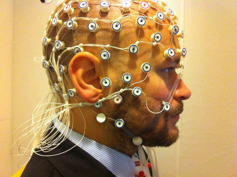 Pasien berbaring di ranjang laboratorium dengan elektroda di kepala, bersiap menjalani perekaman EEG bersama petugas medis.