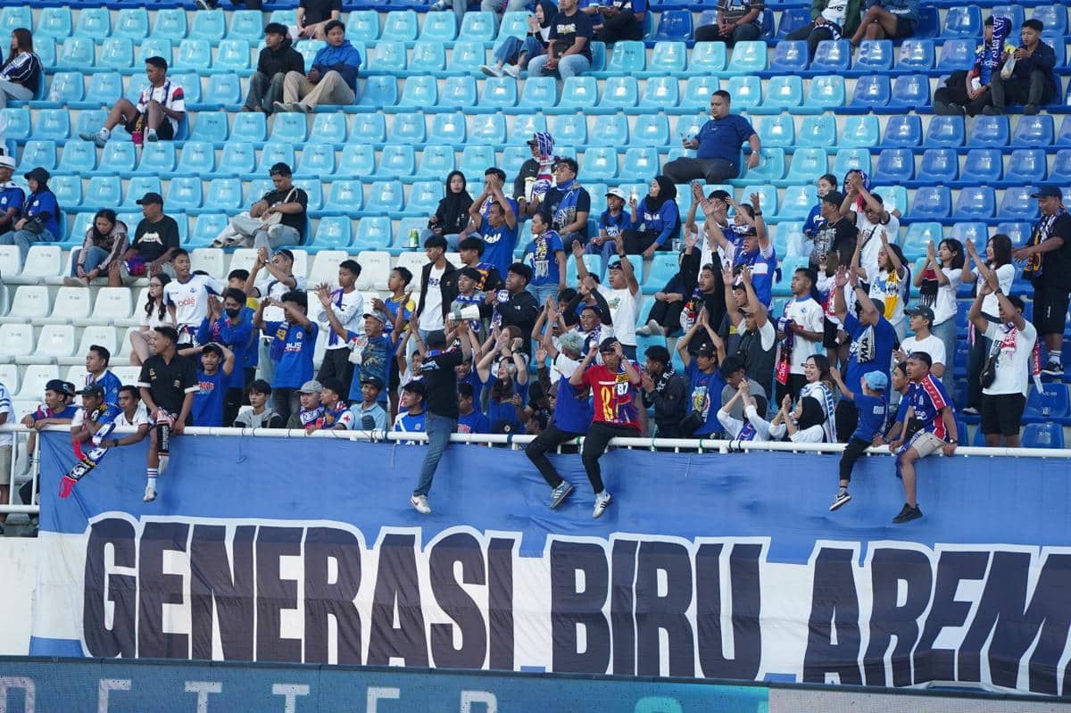 Suporter Aremania berdiri di tribun stadion sambil memberikan dukungan untuk Arema FC dalam suasana pertandingan.