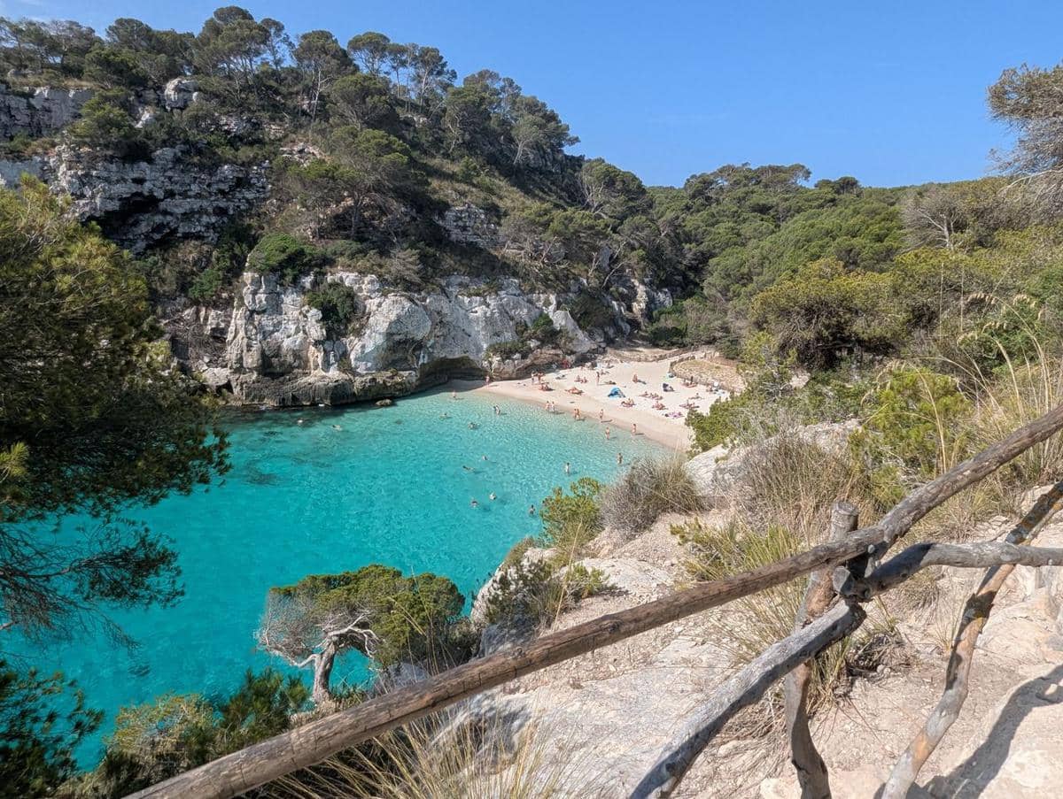Teluk Cala Macarella di Menorca dengan air laut biru jernih, pantai berpasir, dan tebing hijau berbatu di sekelilingnya.