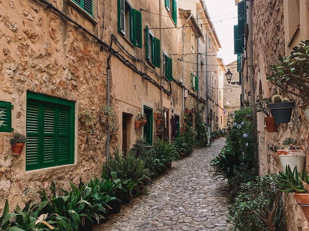 Jalan berbatu di Kota Tua Pollença, Mallorca, diapit bangunan batu bersejarah dan deretan tanaman hijau.