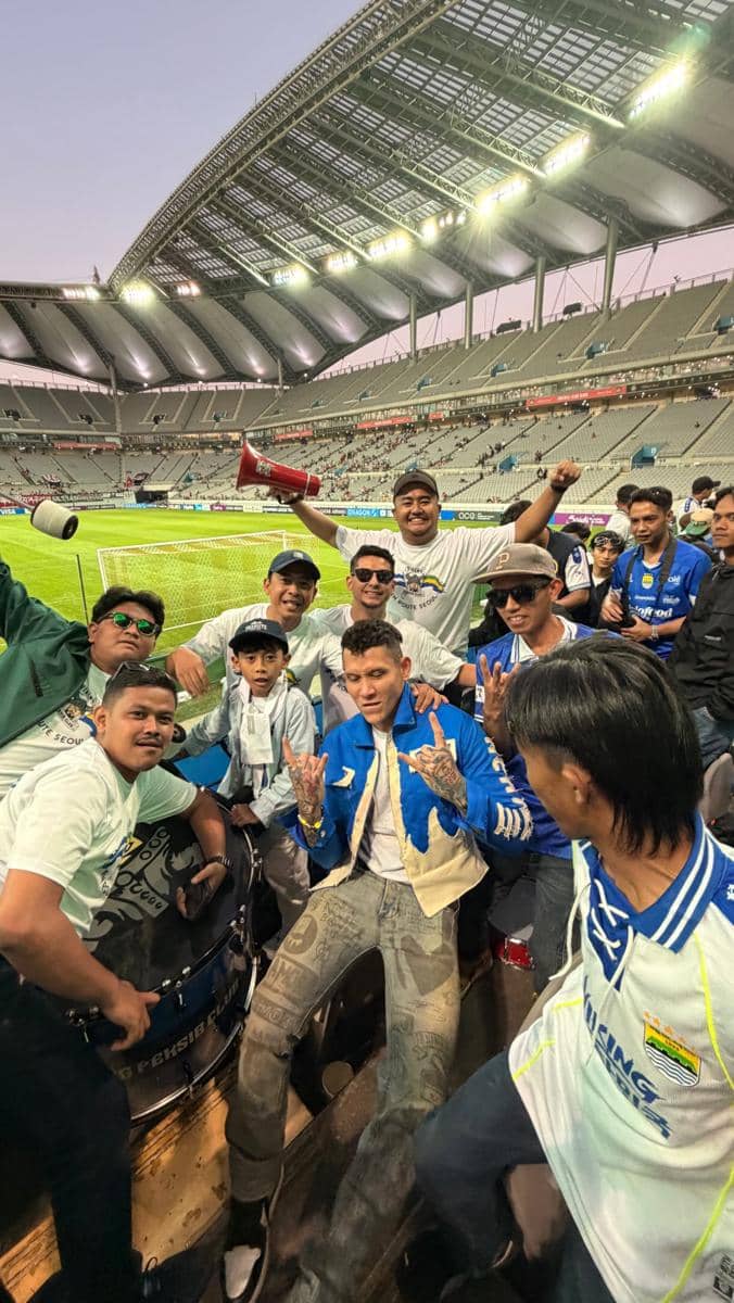 Ganindra Bimo mengenakan jersey Persib sambil berpose di Seoul dengan latar suasana perkotaan Korea Selatan.