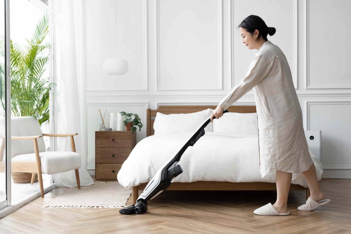 Seorang perempuan membersihkan kamar tidur dengan penyedot debu di dekat tempat tidur dan jendela yang rapi.