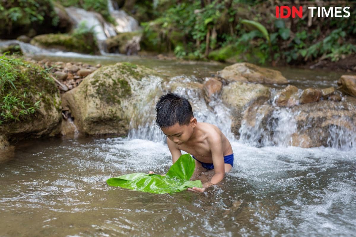 Sejumlah anak bermain ceria di aliran sungai dangkal, menggambarkan air sebagai nikmat yang sering terasa biasa.