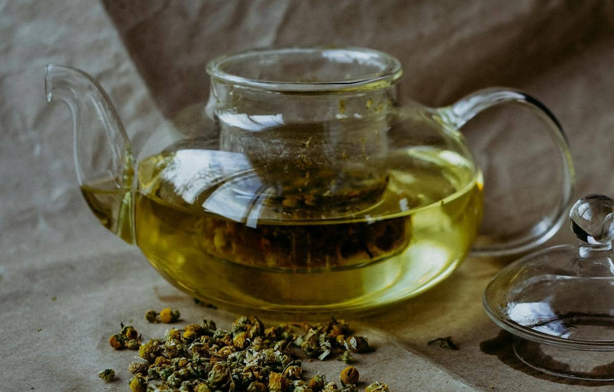 Secangkir teh chamomile hangat berwarna keemasan disajikan bersama bunga chamomile di atas meja kayu.