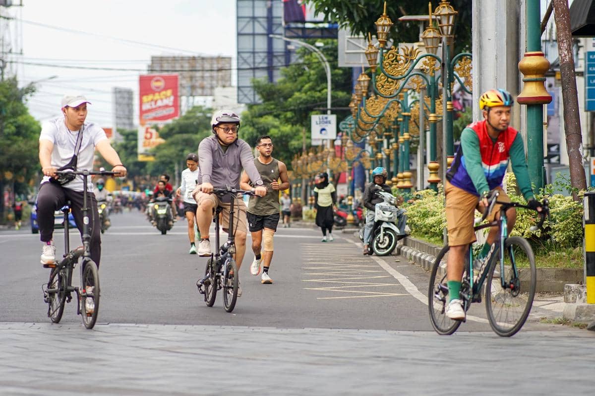 Tiga pesepeda mengayuh sepeda lipat dan sepeda balap bersama warga yang berolahraga lari di jalanan kota.