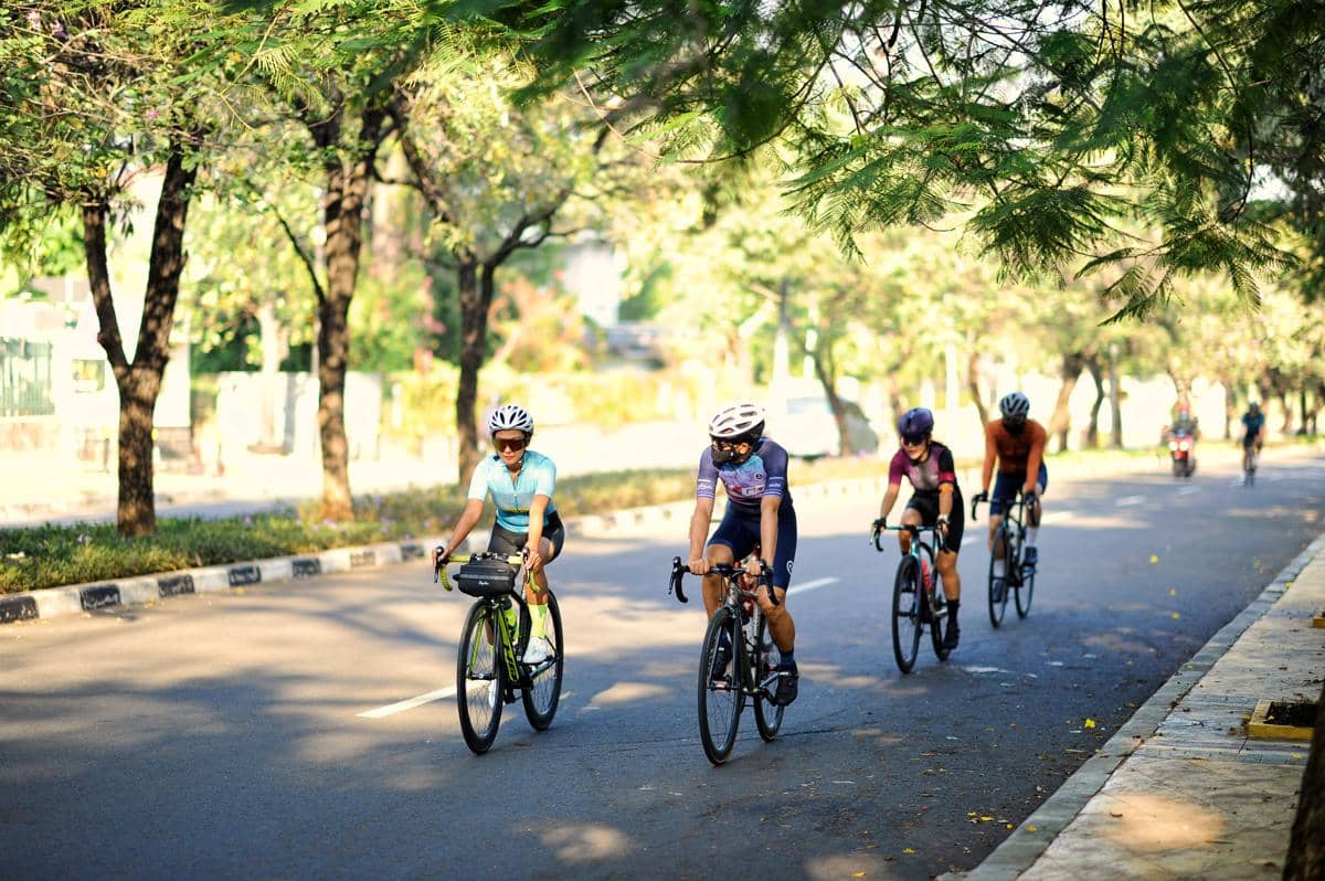 Sekelompok pesepeda menggunakan helm dan jersey mengayuh sepeda balap bersama di jalanan beraspal yang rindang.