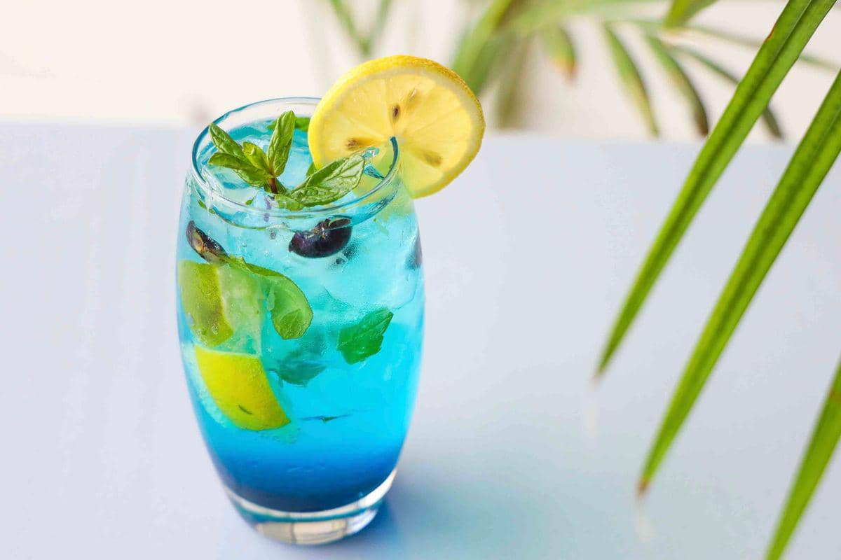 Segelas minuman biru dengan buah leci dan hiasan garam disajikan di atas permukaan berwarna gelap.