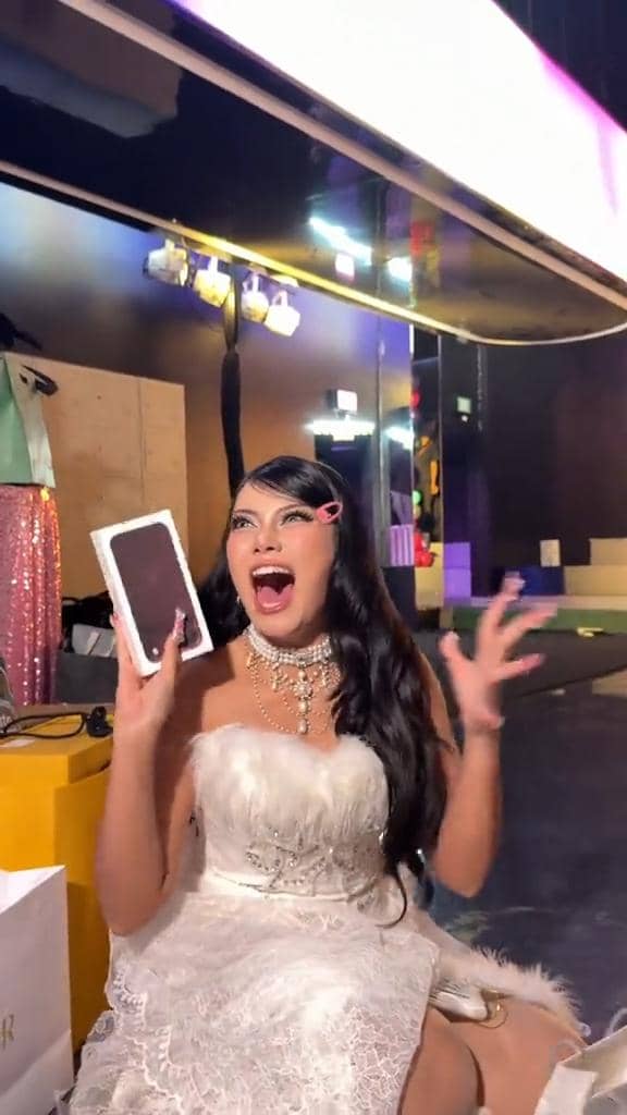 Seorang perempuan berambut panjang memegang iPhone 18 berwarna gelap sambil berbicara di depan kamera.