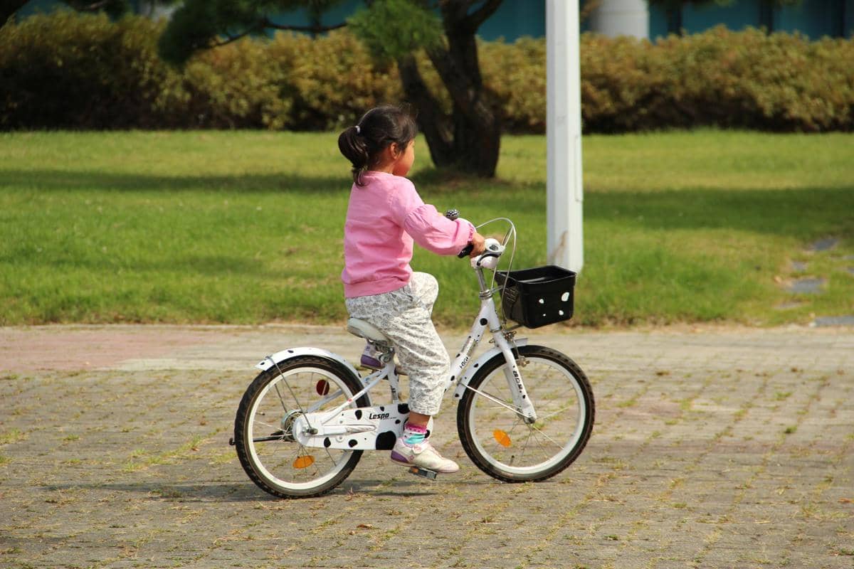 Ilustrasi seorang anak perempuan berbaju merah muda sedang mengayuh pedal sepeda anak berwarna putih di area taman