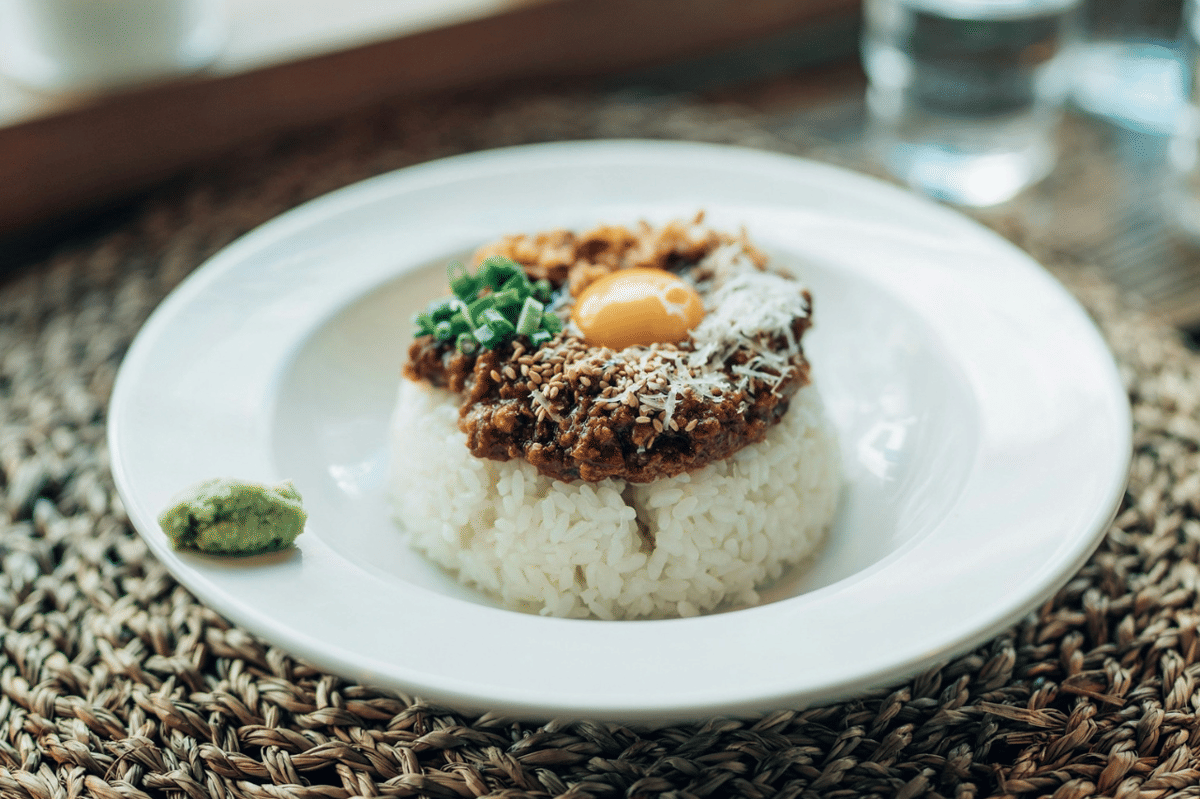 Semangkuk miso hambagu don dengan nasi, patty hamburger, telur mata sapi, dan daun hijau di atas meja.