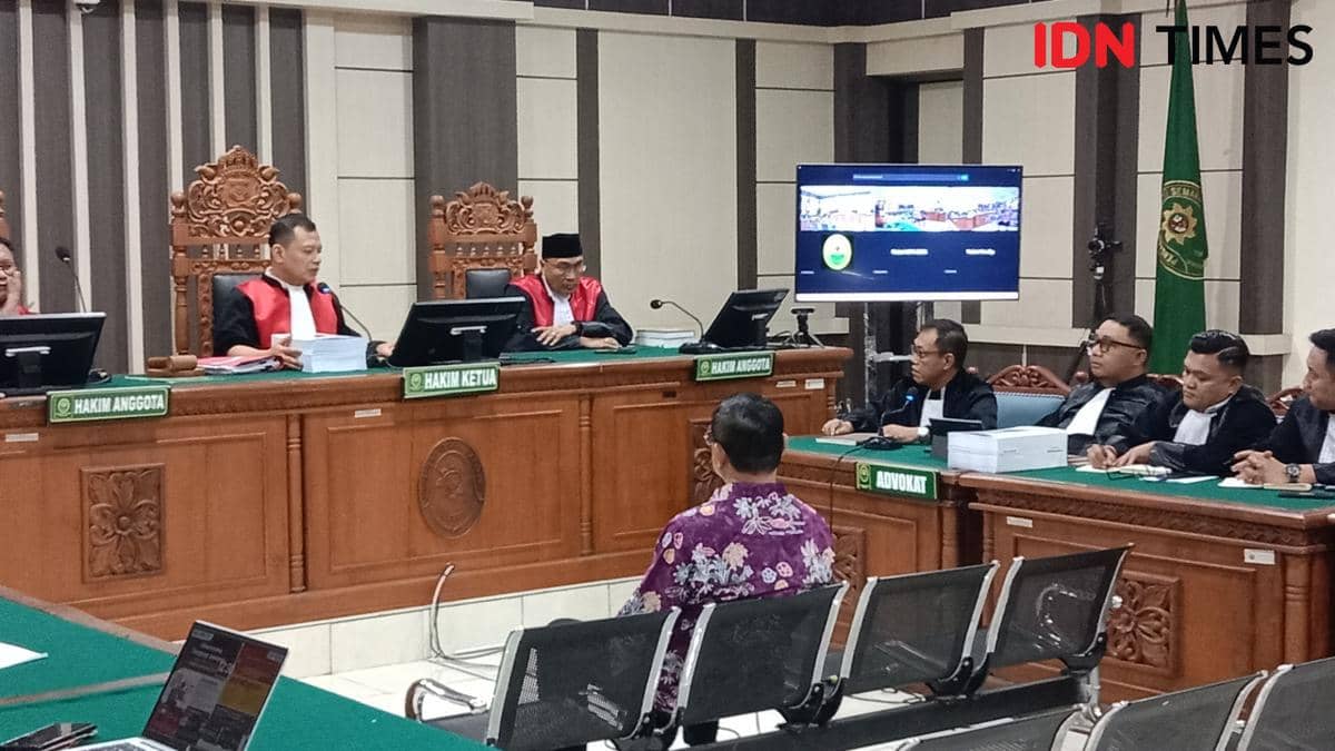 Bupati Pati nonaktif Sudewo duduk di kursi depan ruang sidang dengan mikrofon dan dokumen di hadapannya.