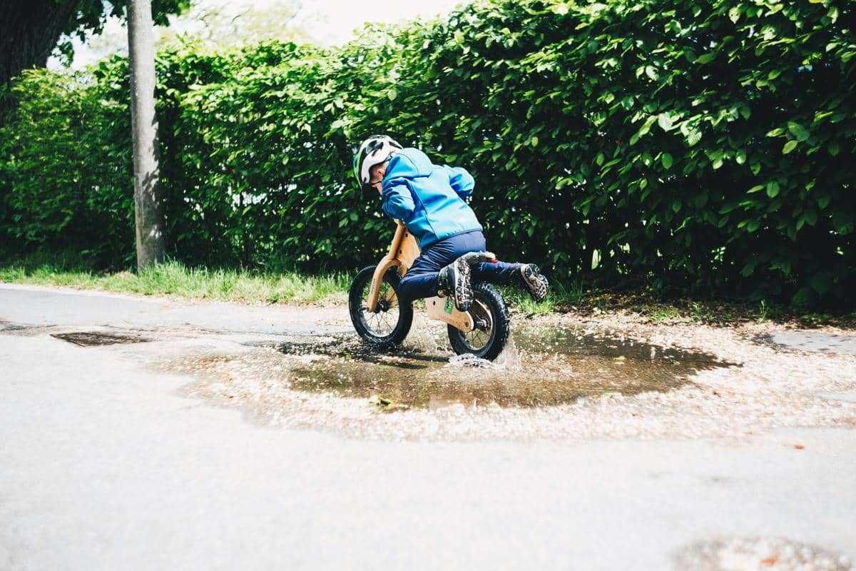 Ilustrasi seorang anak balita mengenakan jaket biru dan helm sedang mengendarai sepeda balance bike kayu menyeberangi genangan air